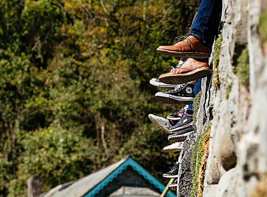 Auf diesem Bild sind mehrere Beine und F&uuml;&szlig;e zu sehen, die &uuml;ber den Rand einer steinernen Mauer baumeln. Die Personen tragen verschiedene Arten von Schuhen.