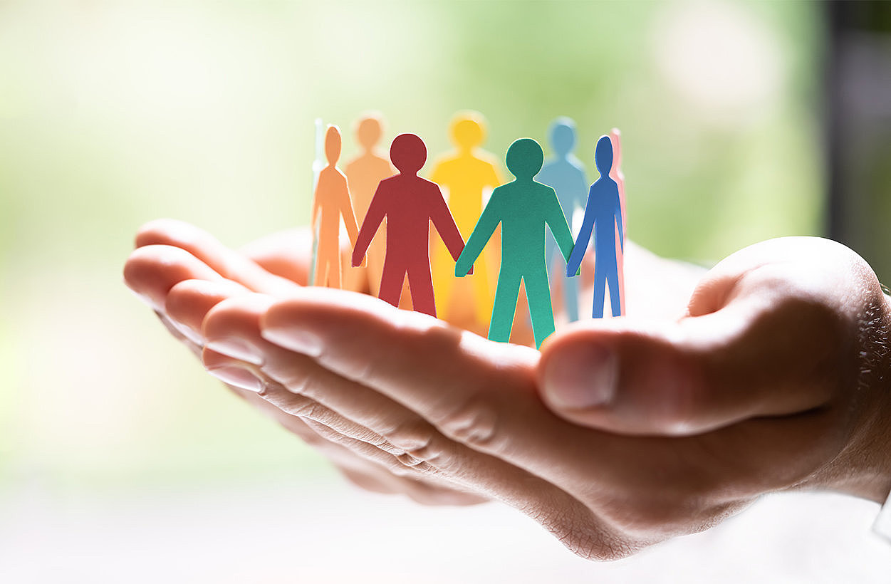 Zwei H&auml;nde halten eine Gruppe von bunten Papierfiguren, die Menschen darstellen, die in einem Kreis stehen. Sie symbolisieren Gemeinschaft, Unterst&uuml;tzung und Zusammenhalt.